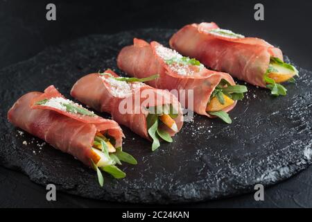 Prosciutto rolls stuffed with arugula and pears on black slate. Stock Photo