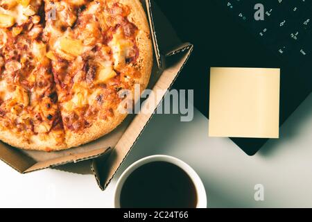 Pizza in box, cup of coffee, laptop and empty sticker with copy space on desk in office. Concept of food or pizza delivery and break on working day. Stock Photo