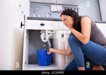 Young Woman Collecting Leakage Water In Bucket At Kitchen Stock Photo