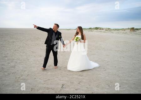 Wedding couple walking on sandy beach. Barefoot bride and groom. Husband pointing at something interesting by hand. Wedding day concept. Stock Photo