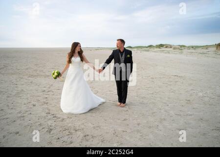 Wedding couple standing and posing on sandy beach. Bride and groom looking to each other and smiling. Barefoot husband. Wedding day concept. Stock Photo