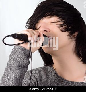 Black hair young guy in emo style is singing in microphone on white background. Stock Photo