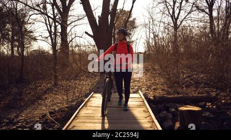 Athlete bicyclist Caucasian woman in sportswear and helmet crosses on foot, leads a mountain bike in her hands across a rural na Stock Photo