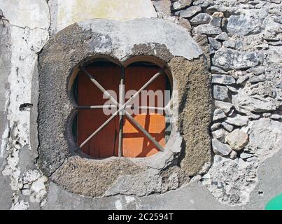 and ancient stone oblong shaped window with crossed bars with an orange shutter set in an old patched repaired grey wall with cr Stock Photo