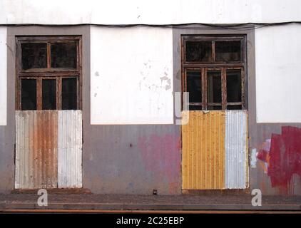 the front of a white and grey painted abandoned house with broken doors and windows boarded up with rusty corrugated iron with s Stock Photo