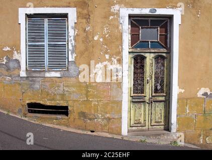 the front of an old abandoned yellow house with blue shuttered windows and locked green wooden door with flaking peeling paint o Stock Photo