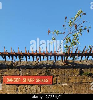 rusty iron sharp spikes on the top of an old stone wall with a red danger sign against a blue sky Stock Photo