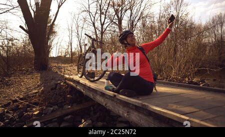 Happy caucasian woman cyclist makes a photo of herself a selfie on the phone while sitting on the bridge over a river in the for Stock Photo
