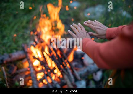 Young woman making fire while camping outdoors, in an alpine wilderness - warming up her hands near the fire Stock Photo