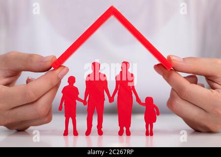 Close-up Of Businesswoman's Hand Protecting Red Family Figures With Red Roof Over White Desk Stock Photo