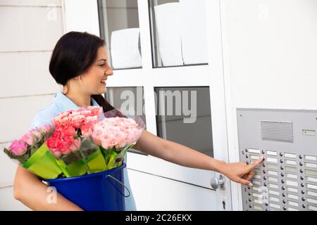 Happy Young Woman Holding Bouquet Pressing Button Of Intercom To Enter Home Stock Photo