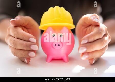 Close-up Of A Person's Hand Protecting Hard Hat And Piggy Bank On White Desk Stock Photo