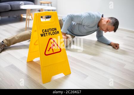 Mature Man Falling On Wet Floor In Front Of Caution Sign At Home Stock Photo