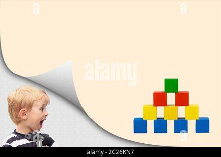 Surprised blond boy in an exposed corner is looking at blank yellow page with colorful wooden cubes toy pyramid. Stock Photo