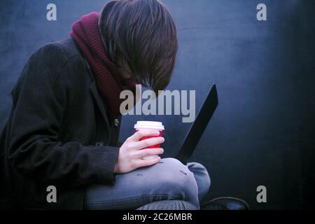The hacker or freelancer person with coffee in hand is using laptop on dark background. Stock Photo