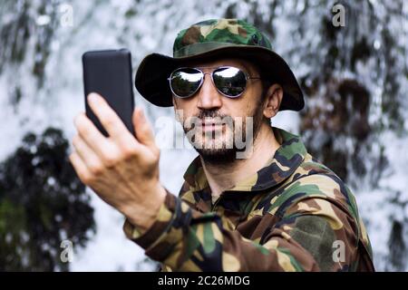 Man in military uniform is taking selfie and using phone in nature, adventure in nature. Stock Photo