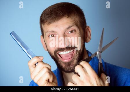 Smiling cheerful male hairdresser is holding a comb and scissors in hands on blue background, haircut for men concept. Stock Photo