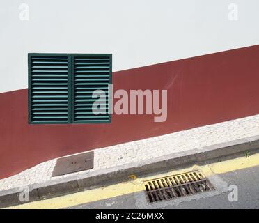 a green shuttered closed window on a red and white house wall on a sloping street with yellow parking line and pavement in typic Stock Photo