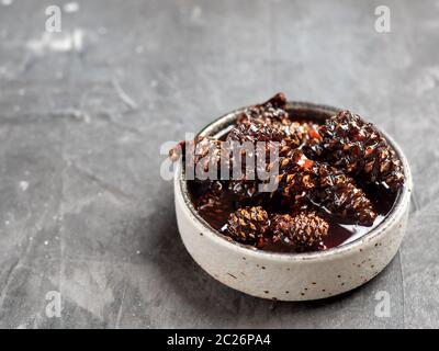 Delicious jam with baby pine cones in small bowl. Traditional Siberian dessert - young pine cones jam on gray textured background. Stock Photo