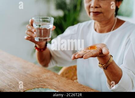 Senior Asian woman with a cup of water and pill taking remedy in cozy room at home. Old female taking medicine Stock Photo