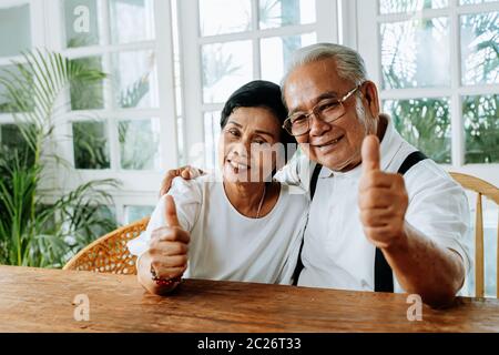 Cheerful Asian man and woman hugging each other and showing thumb up gesture while sitting at table in cozy room at home Stock Photo