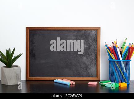 empty black chalk board, multi-colored pencils in a blue metal stand on a black table, concept back to school Stock Photo