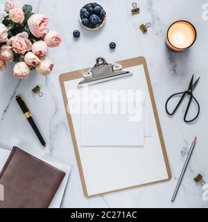 Wedding mock up. Flat lay with blank greeting card, envelope. Clipboard, flowers, scented candle on white marble. Feminine home office mock up with co Stock Photo