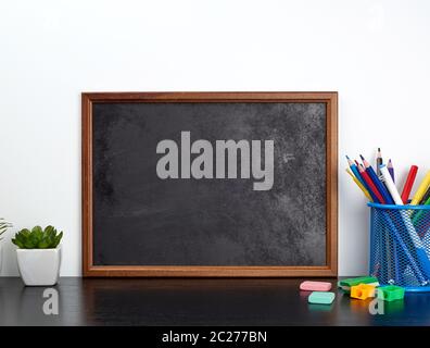 empty black chalk board, multi-colored pencils in a blue metal stand on a black table, concept back to school Stock Photo