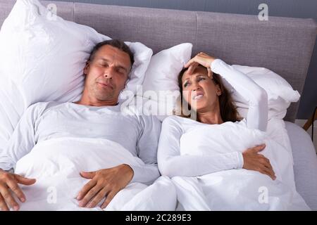 Mature Angry Woman Having Headache While Lying With Snoring Husband In Bed Stock Photo