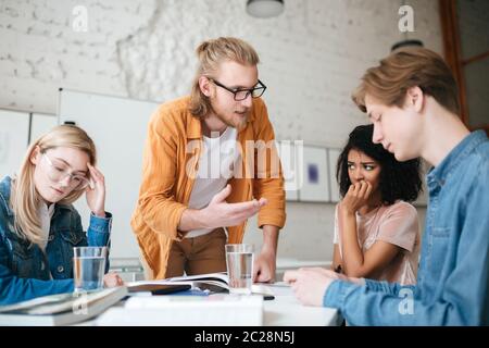 Portrait of teacher with blond hair and beard leaning on table and emotionally explaining something to students. Group of upset people sitting at the Stock Photo
