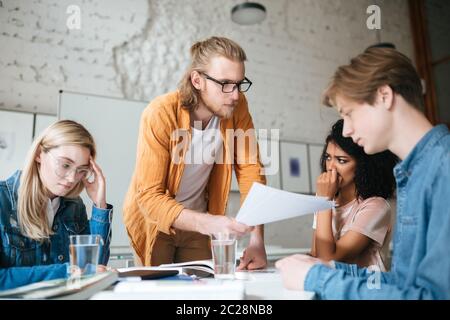 Portrait of young serious teacher in glasses leaning on table and explaining something to students with tests in hands. Group of upset students sittin Stock Photo