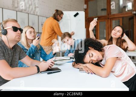 Young teacher writing on board while students sitting at the table and aren't listening him during lesson. Group of students making photos,listening m Stock Photo