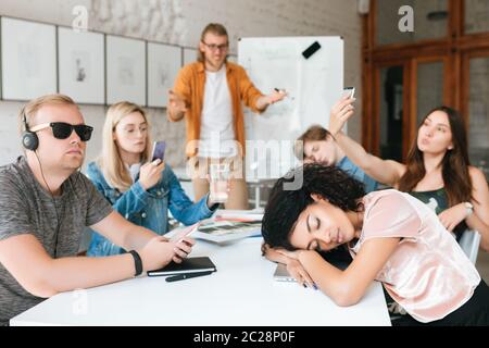 Teacher standing near board and amazedly looking at students. Group of young students making photos, listening music and sleeping during lesson. Stude Stock Photo
