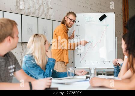 Young smiling teacher standing near board and happily looking at students while explaining them diagram. Group of young students listening teacher dur Stock Photo