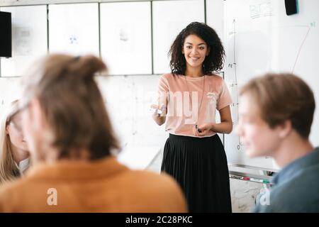 Portrait of beautiful African American lady with dark curly hair standing near board and giving presentation to colleagues in office. Young smiling bu Stock Photo