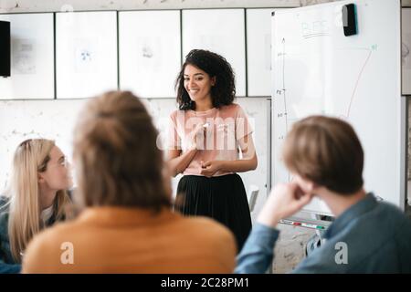 Portrait of cheerful African American lady with dark curly hair standing near board and happily speaking with colleagues in office. Young beautiful bu Stock Photo