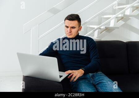 Handsome pensive young man using laptop, sitting on cozy sofa . Black and white interior. At home. Stock Photo