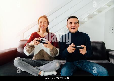 Pretty girl and a guy playing video games online, sitting on couch together, dressed in warm casual clothes. At home. Stock Photo