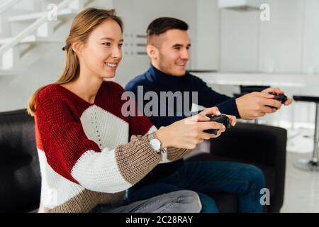 Beautiful young couple playing video games using joysticks, sitting on couch at home. Dressed in warm casual clothes. Side view. Stock Photo