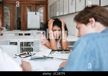 Portrait of upset African American girl with dark curly hair sitting in library with books on table and and holding hands by head. Group of young tire Stock Photo