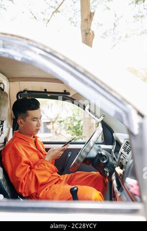 Young Asian man working in house moving service company sitting relaxed in van using his smart phone, vertical shot Stock Photo