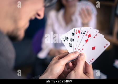 Friends are playing cards together at home. Man is holding cards in her hands, woman in the blurry background. Stock Photo