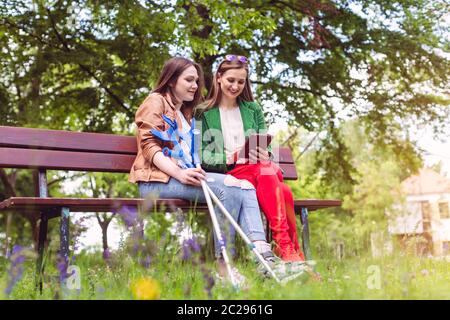 Two friends in a park, one with a broken feet and crutches reading a book Stock Photo