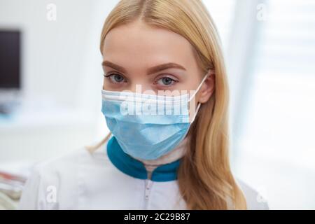 Close up of a blond haired female doctor wearing medical mask, looking to the camera seriously. Female practitioner posing at the hospital. Medical wo Stock Photo