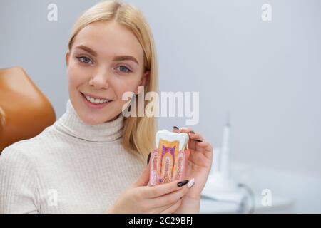 Beautiful young woman smiling to the camera, sitting at dentist office, copy space. Attractive female patient holding tooth mold, posing at dental cli Stock Photo