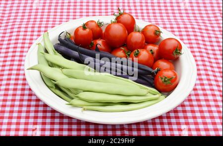 Green yin yang beans, purple French beans and juicy red cherry tomatoes in a round white dish on a red gingham tablecloth Stock Photo