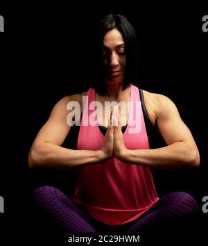 young beautiful athletic girl in a pink top sits with crossed legs in a lotus position, hands makes a gesture of namaste with joined hands in front of Stock Photo