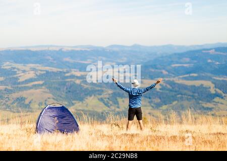 Young tourist man admire beautiful mountains view with arms up near tent, camping place Stock Photo