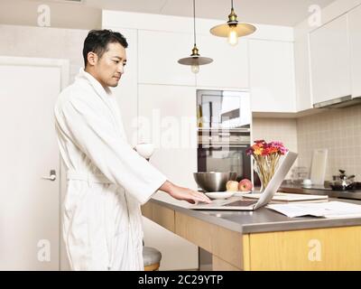 young asian business man in bathrobe working from home standing by kitchen counter holding a cup of coffee using laptop computer Stock Photo