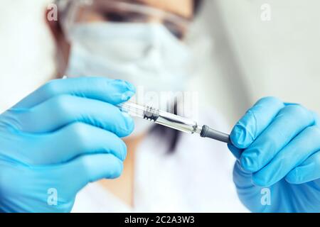 Hands in medical gloves holds a syringe. Female doctor in protecive glasses, face mask and gloves holds syringe in her hands. Stock Photo